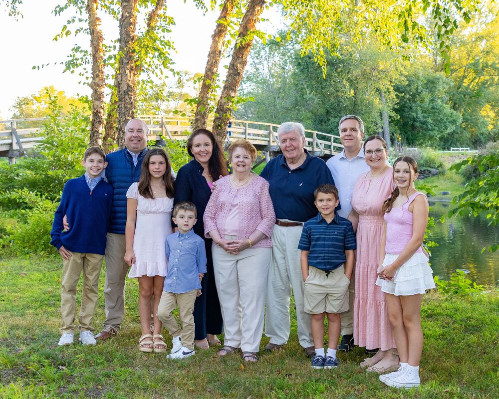 A multi-generational family group poses together outdoors on a summer evening by Concord River with trees in the background in Concord, MA.