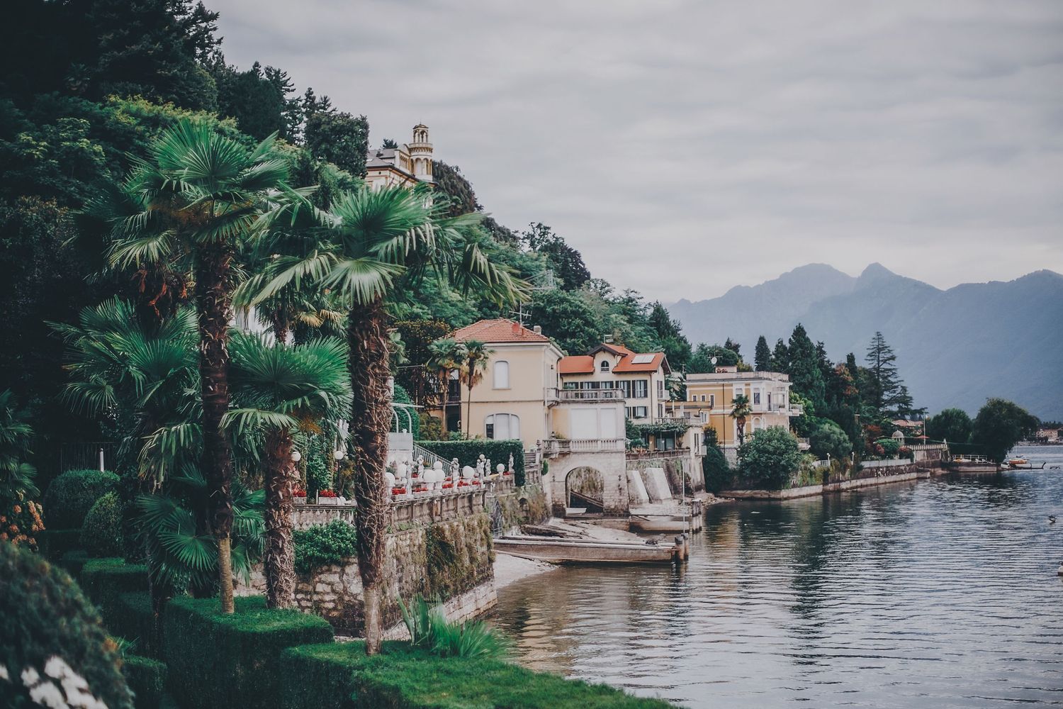 Italian lakeside villas with stone architecture and lush greenery overlook tranquil waters on a cloudy day.