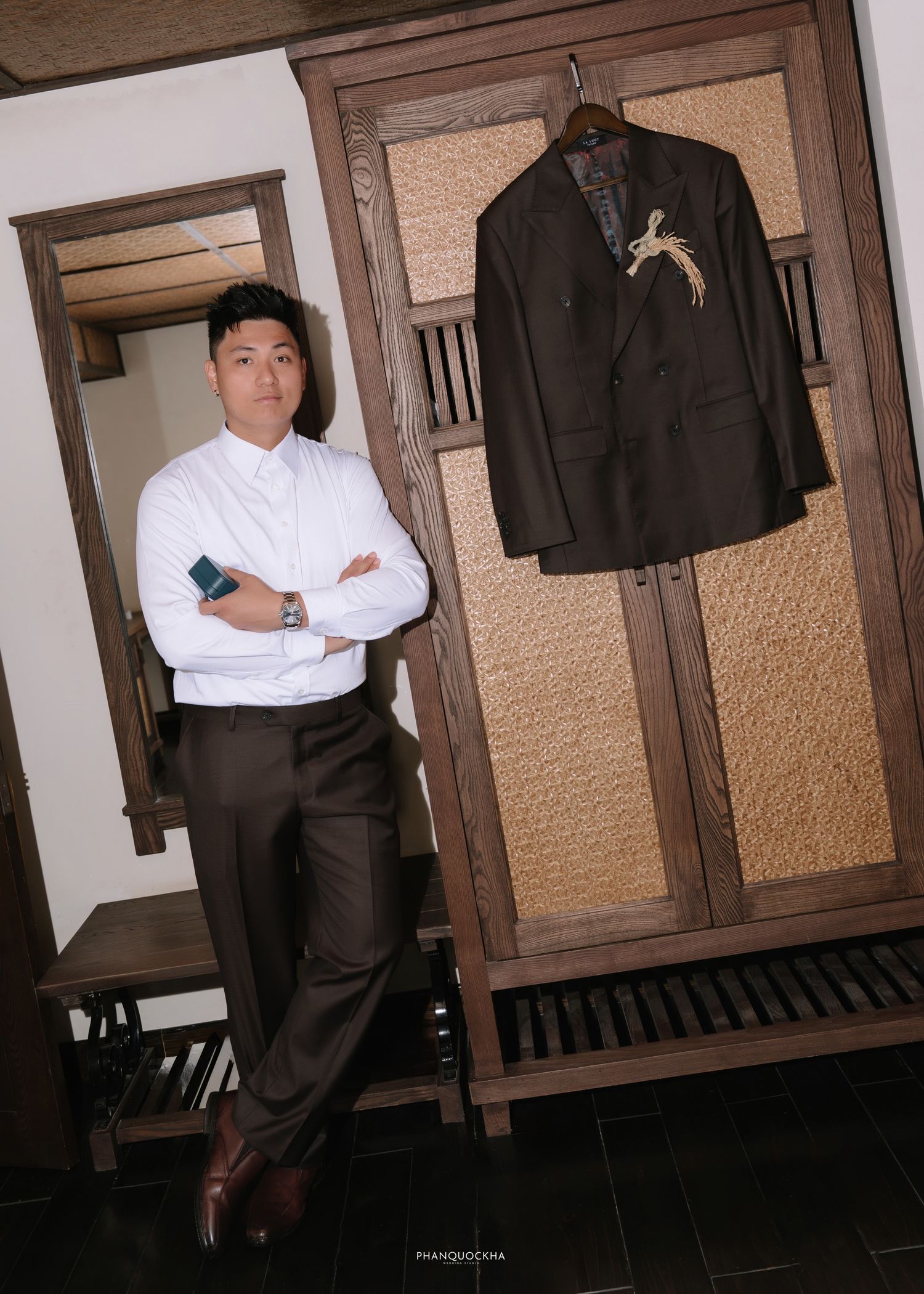 Groom in white shirt posing beside a dark wooden wardrobe with his dark suit jacket hanging inside.