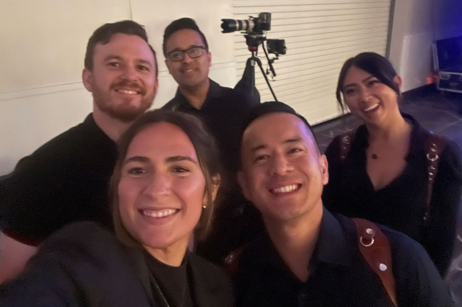 Five smiling event professionals pose for a selfie in a dimly lit venue with camera equipment behind them.