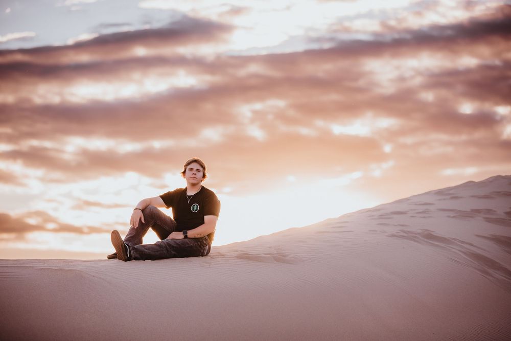 A person lies atop a sand dune at sunset, silhouetted against a dramatic pink and orange cloudy sky.
