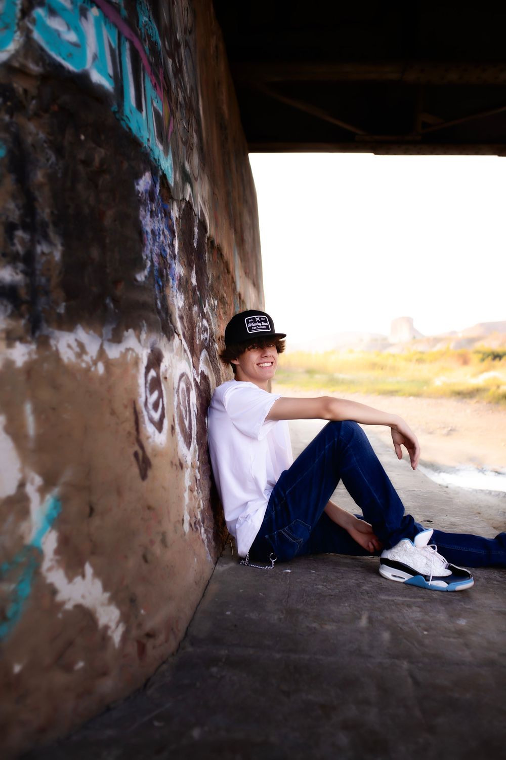 A person in dark clothes and hat sitting under a tunnel or bridge with graffiti-covered walls during sunset.