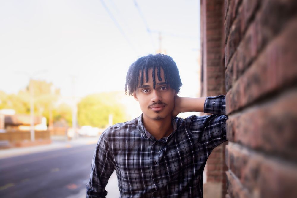 Person in plaid shirt and glasses poses against brick wall in outdoor portrait series.