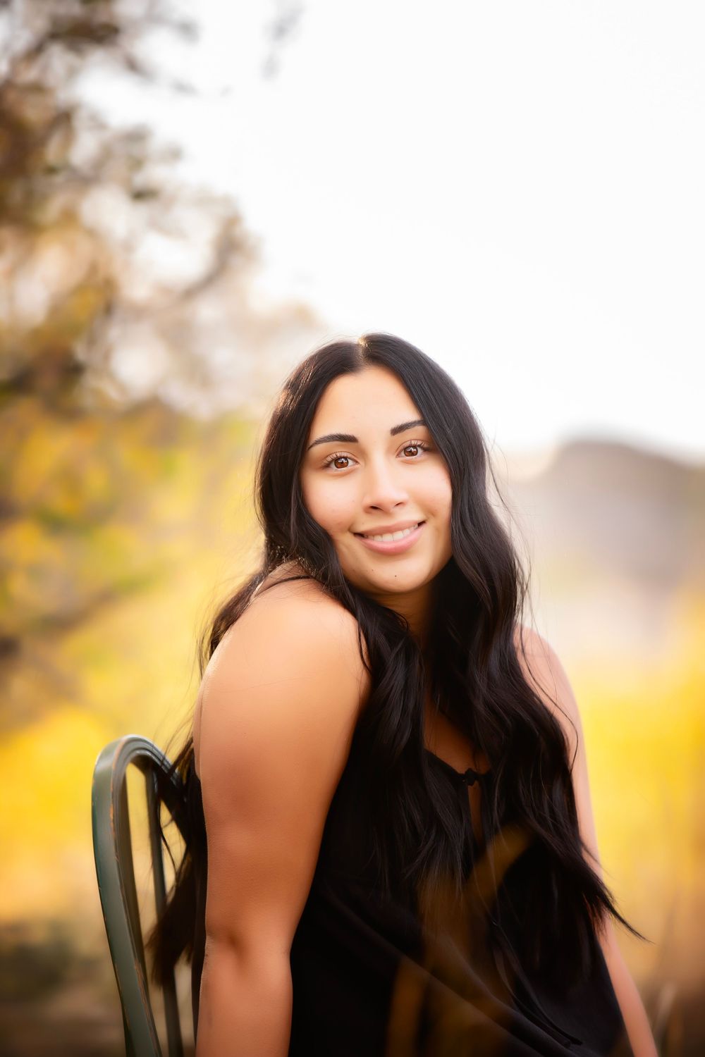 Portrait of someone with long dark hair sitting in a chair outdoors with autumn colors in the background.