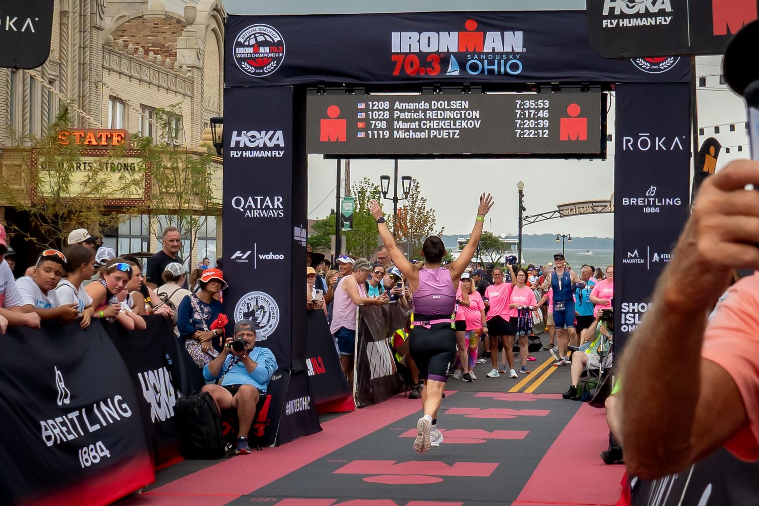 Athletes running across red carpet path at an Ironman triathlon event setup with crowd and barriers along the course.