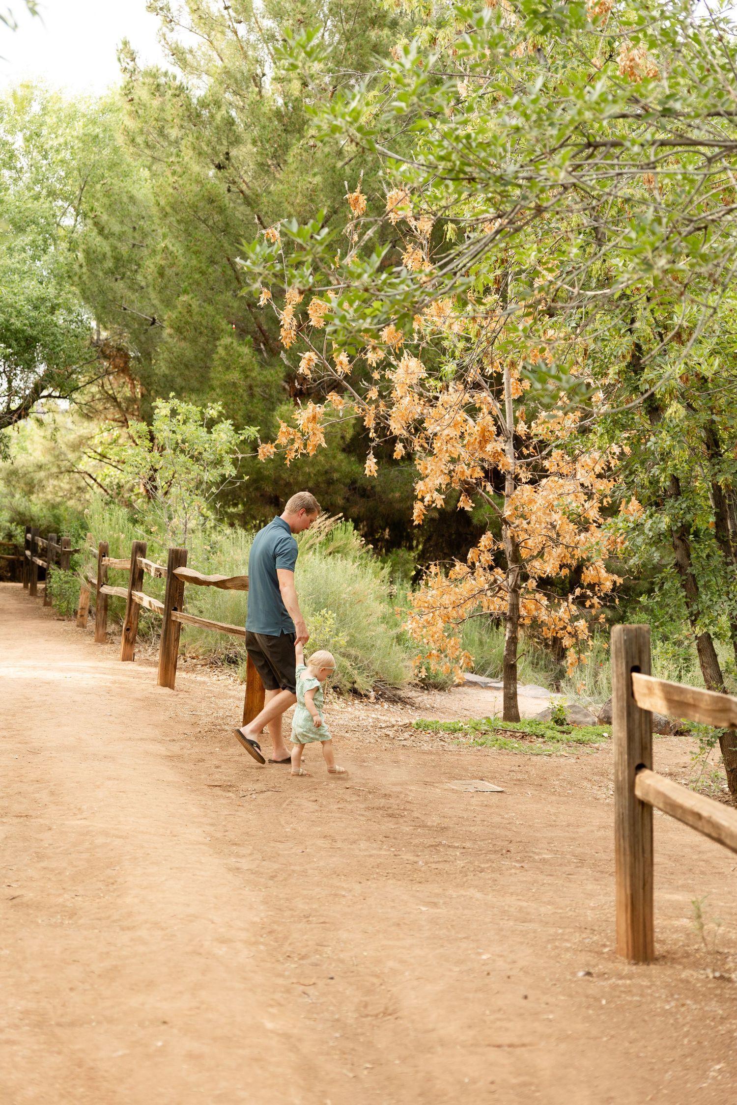 a dad walking with a 1 year old child down a dirt path with trees in the background