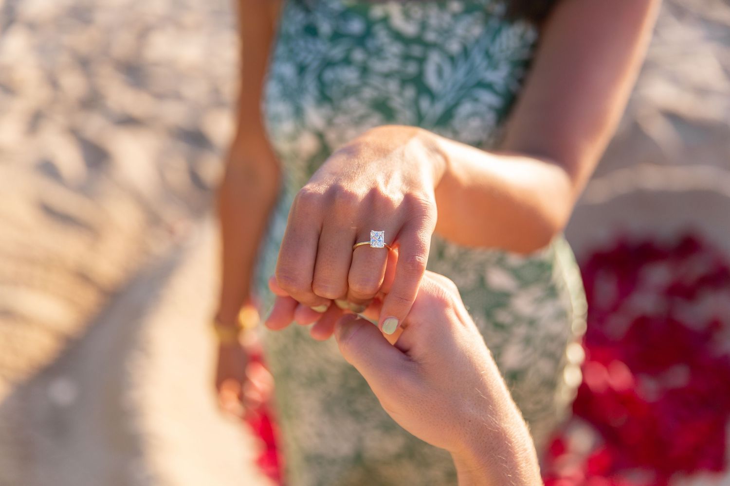Close up view of a proposal wearing a diamond ring, a delicate pearl bracelet with silver chain highlights against blurred sand and roses.