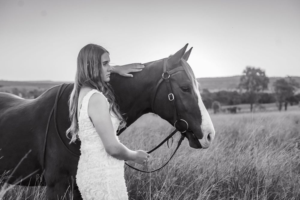 A woman in a white lace dress stands lovingly beside a dark horse in a grassy field in black and white.