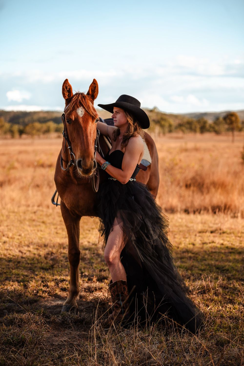 Romantic sunset photoshoot in golden field with woman in black lace gown posing with rustic brown horse.