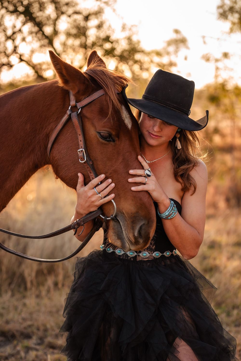 Person in black dress and cowboy hat affectionately interacting with chestnut horse in warm evening light.
