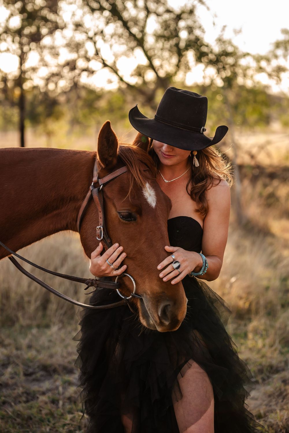 Woman in black dress and cowboy hat shares tender moments with brown horse in golden sunset light.
