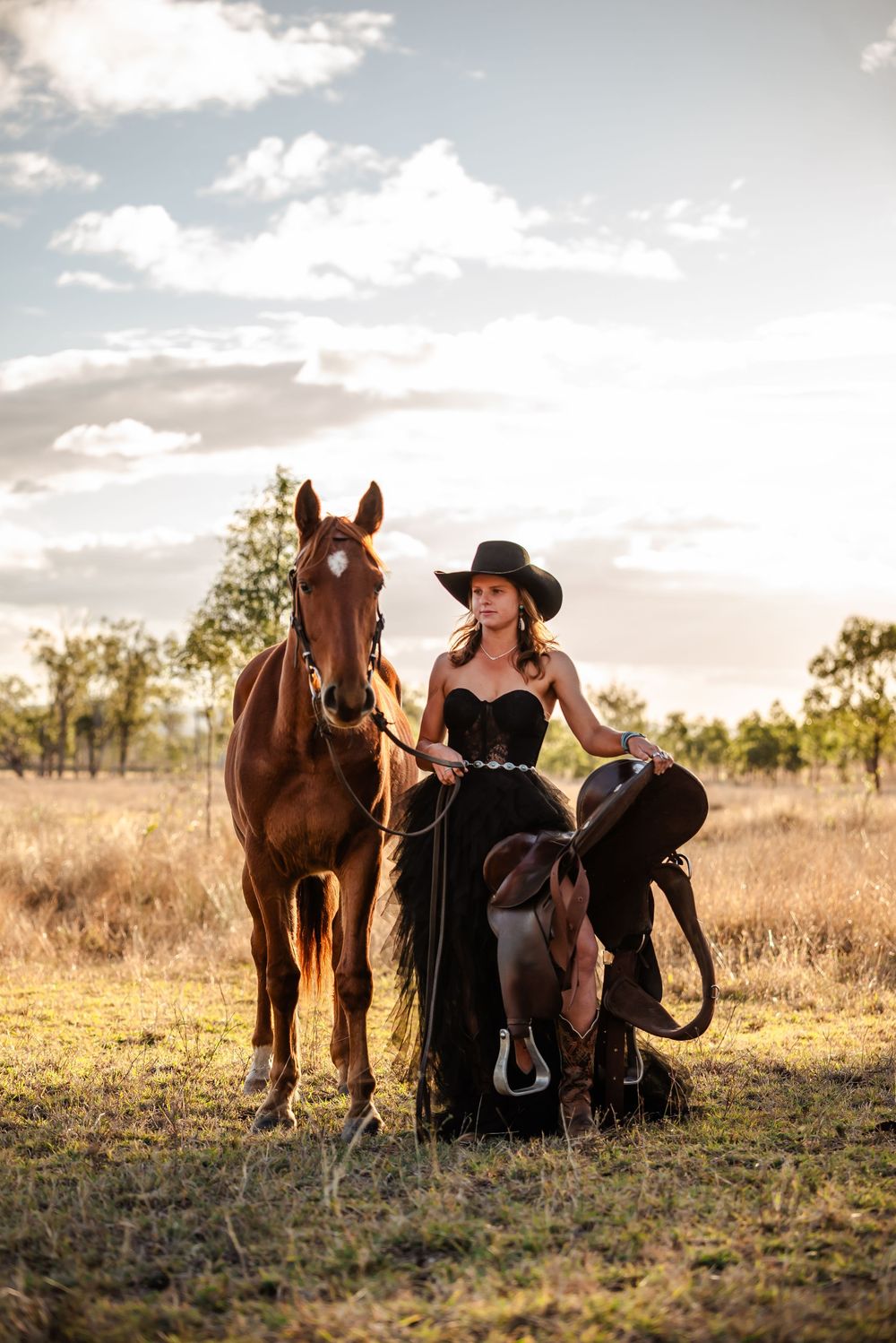 A cowgirl in black attire sits with her horse in a golden field at sunset, creating a serene western scene.