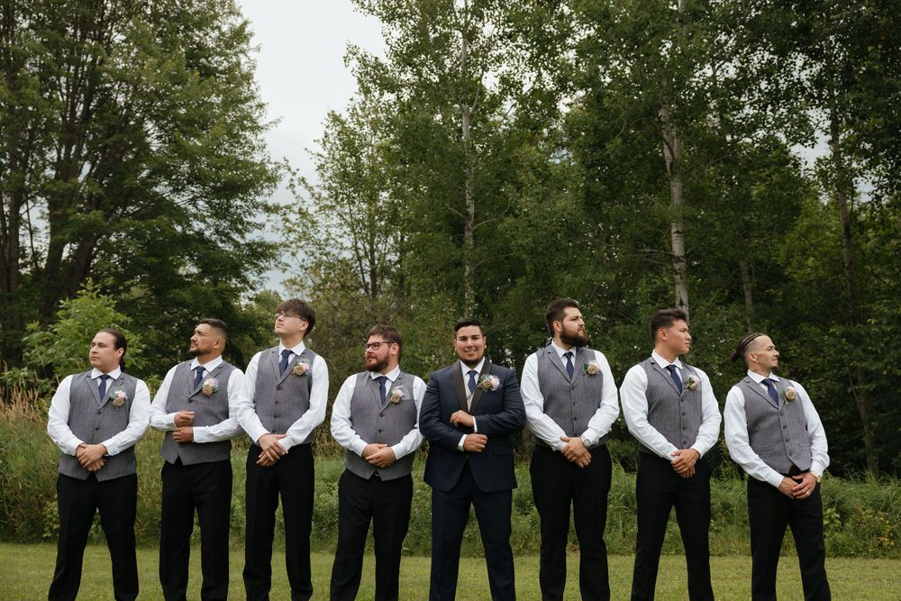 Wedding party in gray vests stands in line against green trees and grass outdoors.