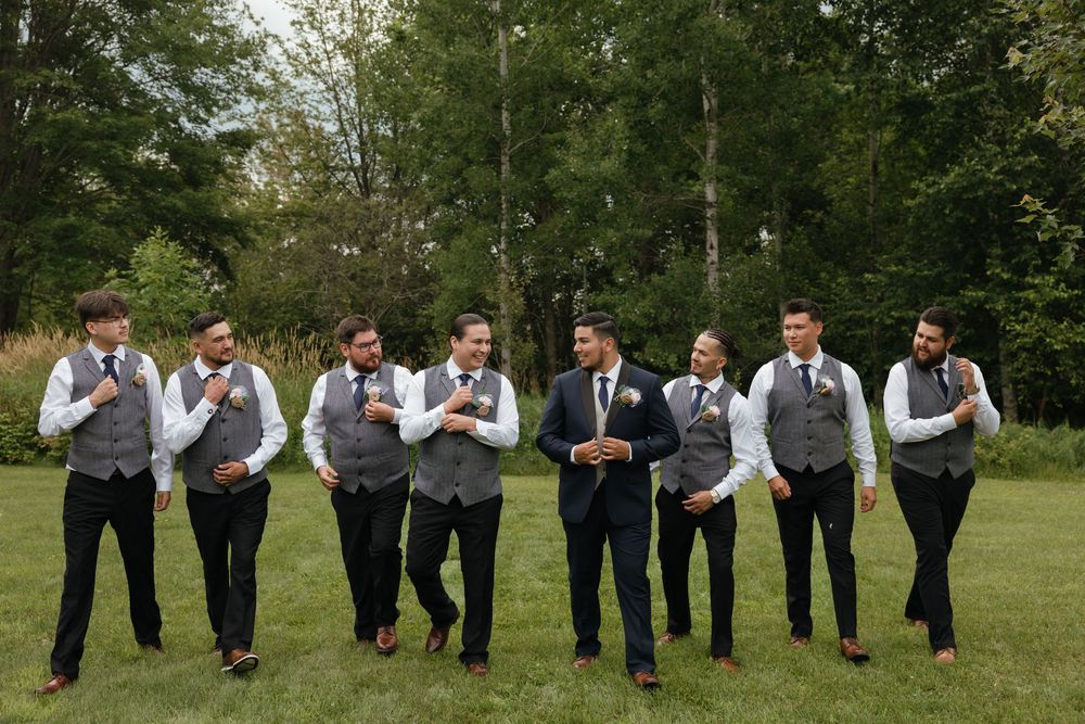 A wedding party in dark suits stands on grass and performs synchronized jumping poses in a series of fun photos.