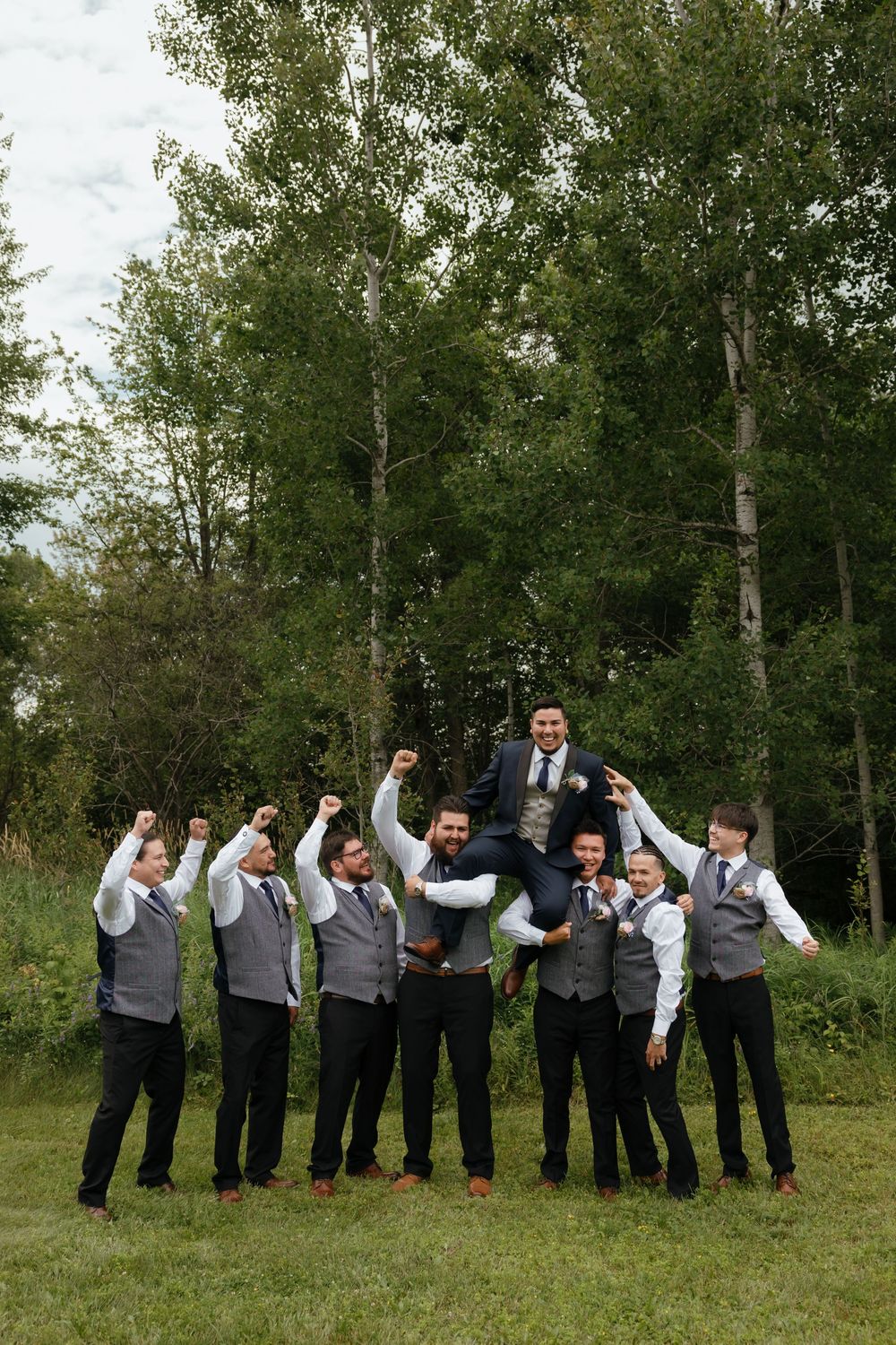Groomsmen in gray vests perform a synchronized jumping pose on grass with trees in the background.