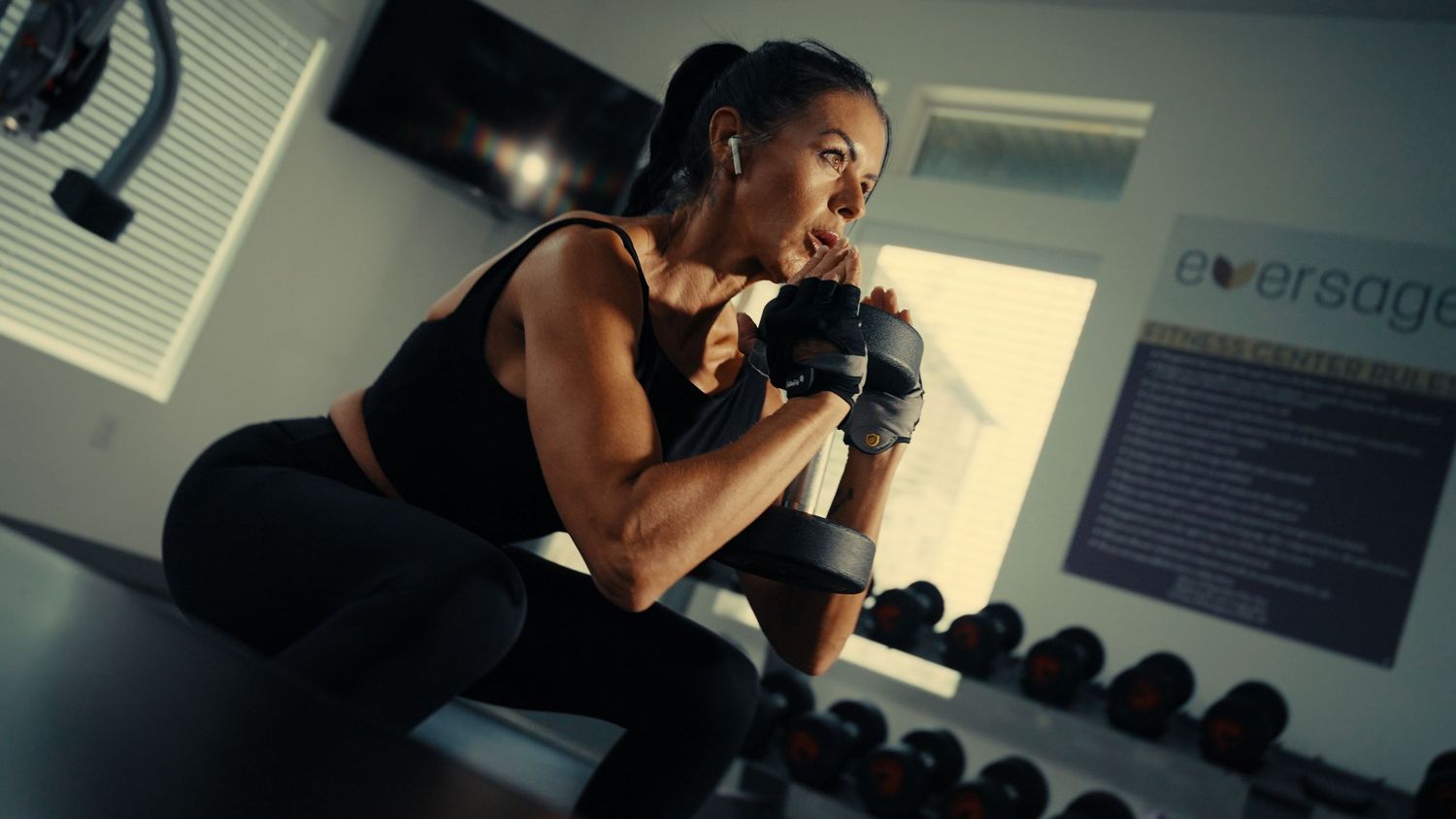 Athletic person in black workout attire performs dumbbell exercises at a fitness gym with weights visible in background.
