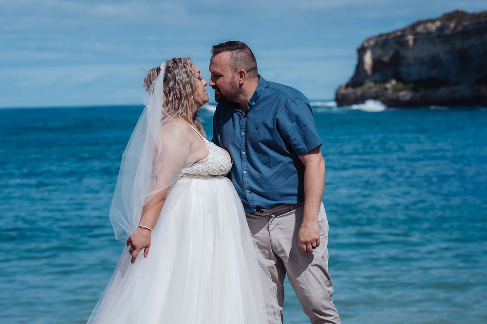 Couple in wedding attire shares romantic moment on beautiful tropical beach with turquoise waters and rocky coastline.