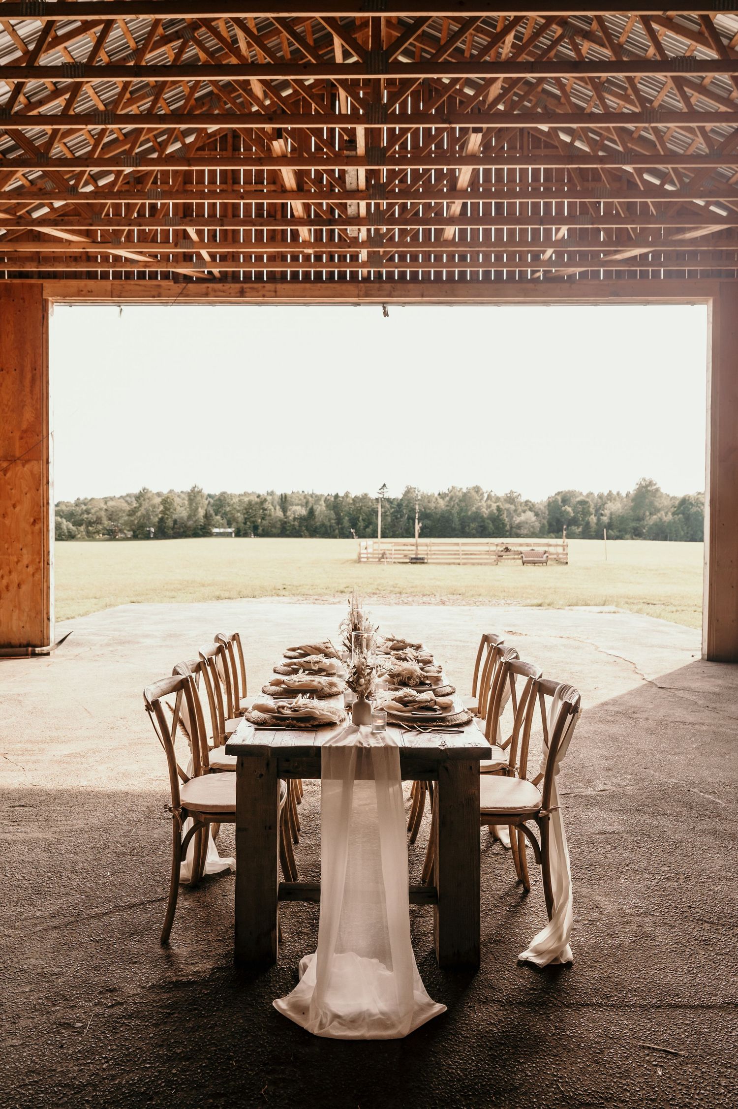 Six round dining tables with wooden chairs are elegantly set inside a rustic barn venue with exposed wooden beams.