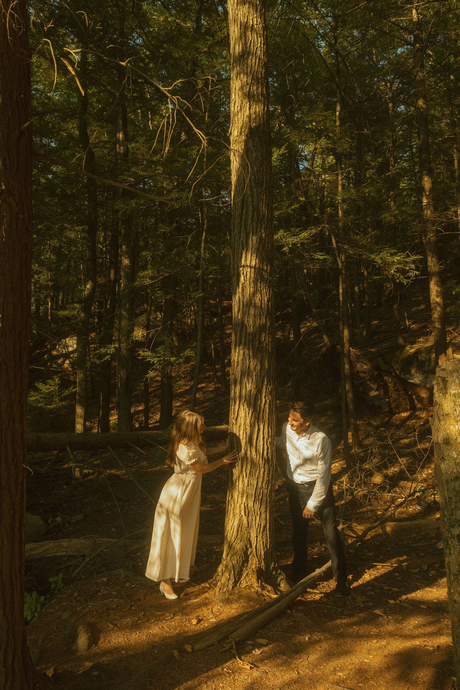 Two people in white outfits pose artistically against tall trees in a sunlit forest.