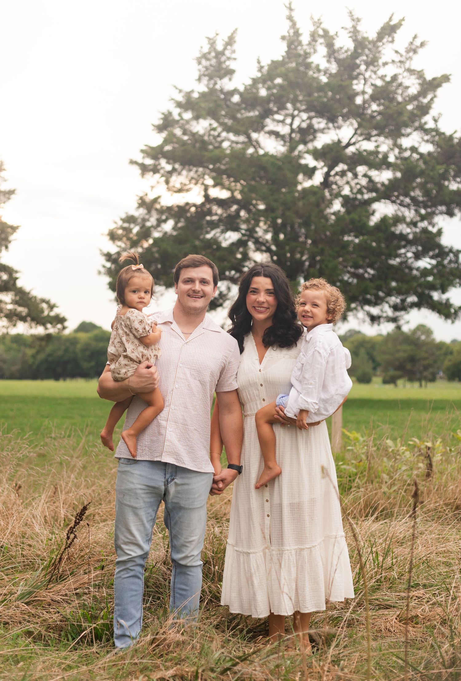 Young family of four enjoying outdoor lifestyle photos in a wooded field. Parents hold their toddler and baby girl while smiling.