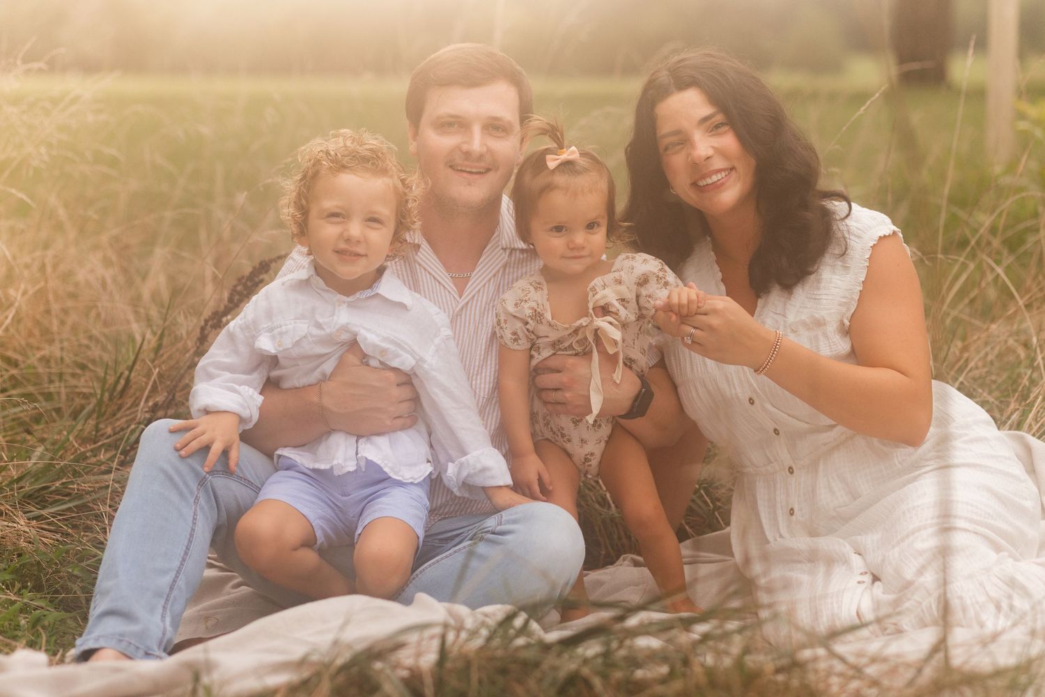 Young family of four enjoying outdoor lifestyle photos in a wooded field. Parents hold their toddler and baby girl while smiling.