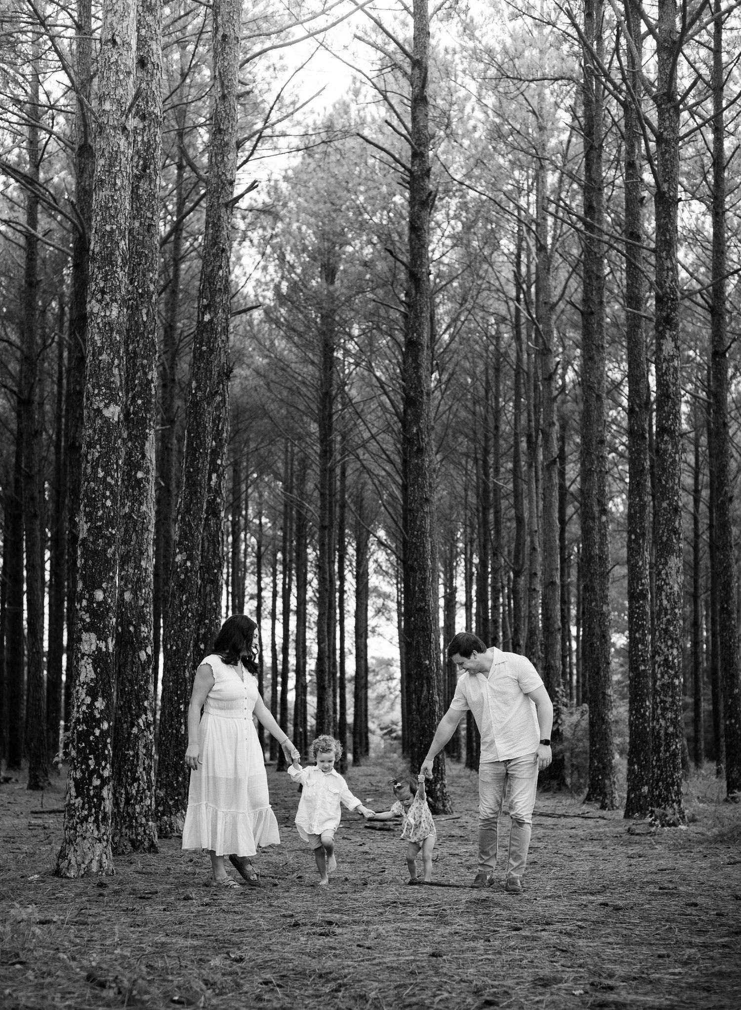 Young family of four enjoying outdoor lifestyle photos in a wooded field. Parents hold their toddler and baby girl while smiling.