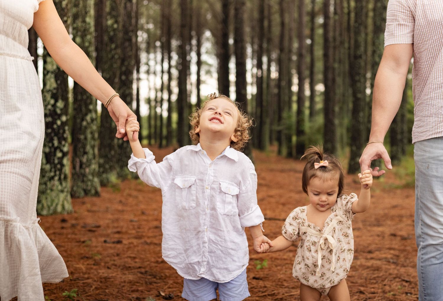 Young family of four enjoying outdoor lifestyle photos in a wooded field. Parents hold their toddler and baby girl while smiling.