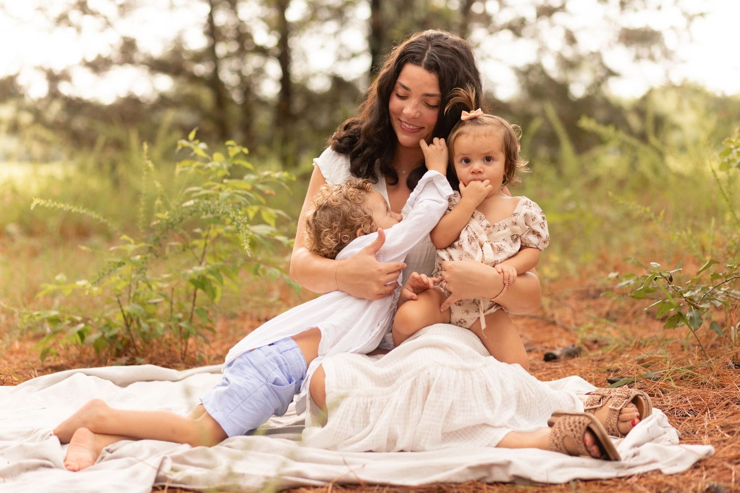 Young family of four enjoying outdoor lifestyle photos in a wooded field. Parents hold their toddler and baby girl while smiling.