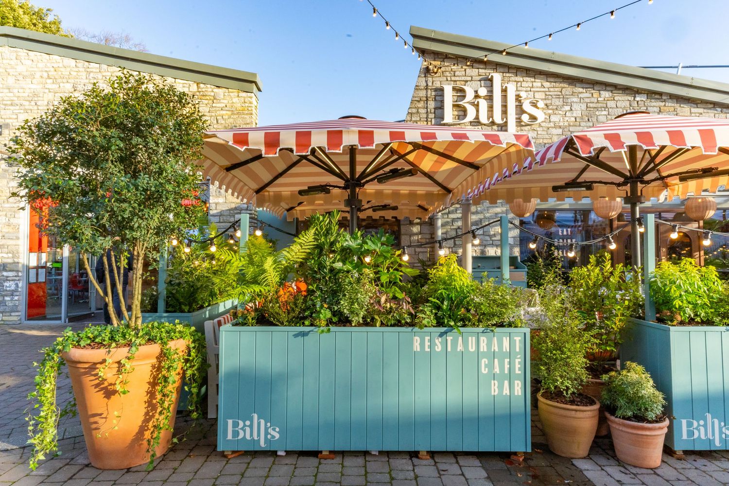 A row of colorful potted plants and outdoor decor line the exterior wall of Bills restaurant with turquoise accents.