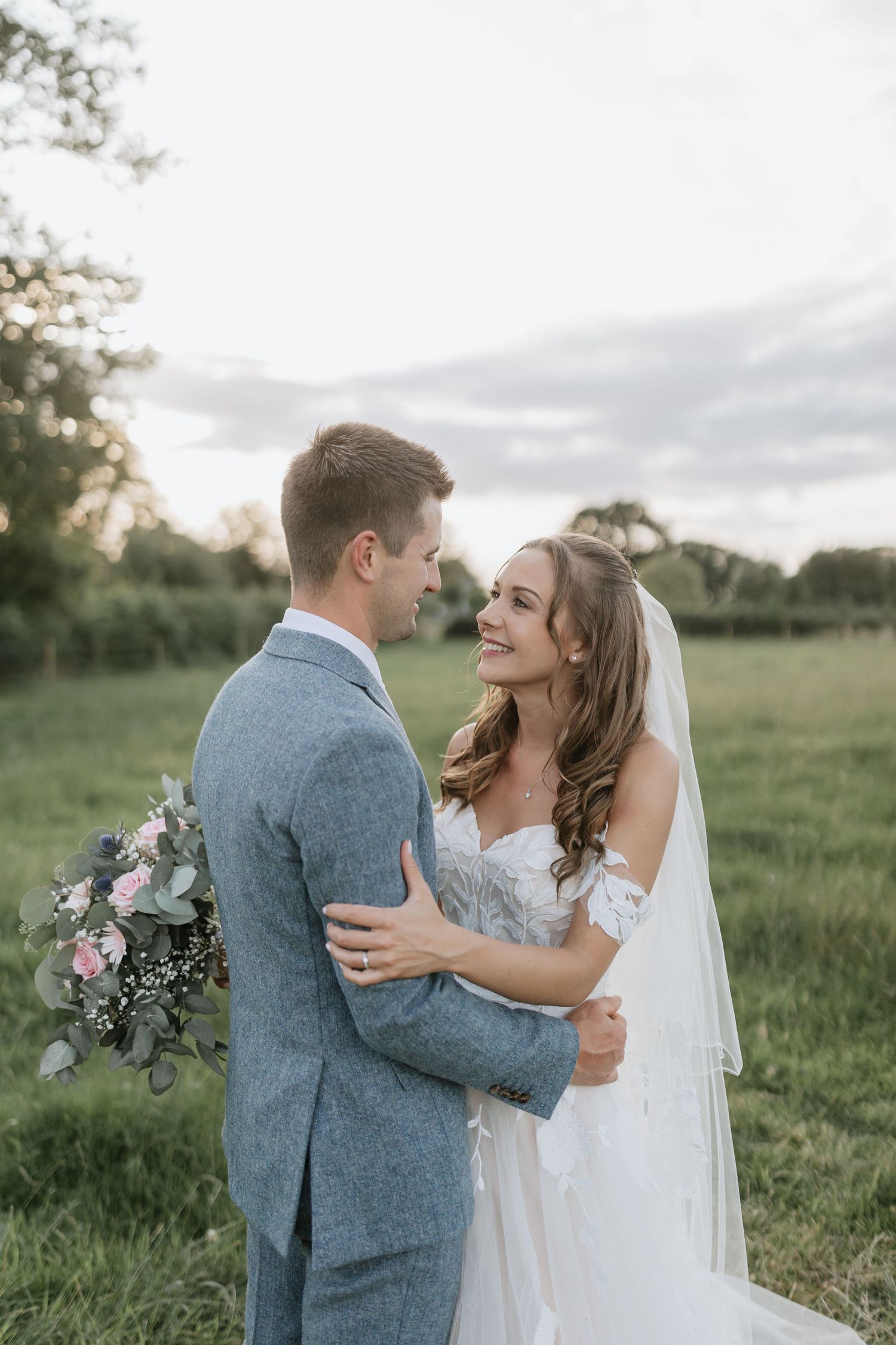 A couple in wedding attire share intimate moments outdoors in a pastoral field as sunlight gently filters in.