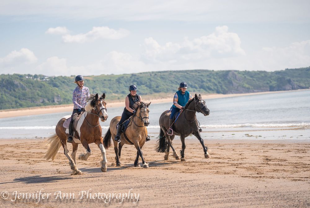 Three riders gallop horseback at Oxwich Beach along a scenic sandy beach with cliffs and ocean in the background.