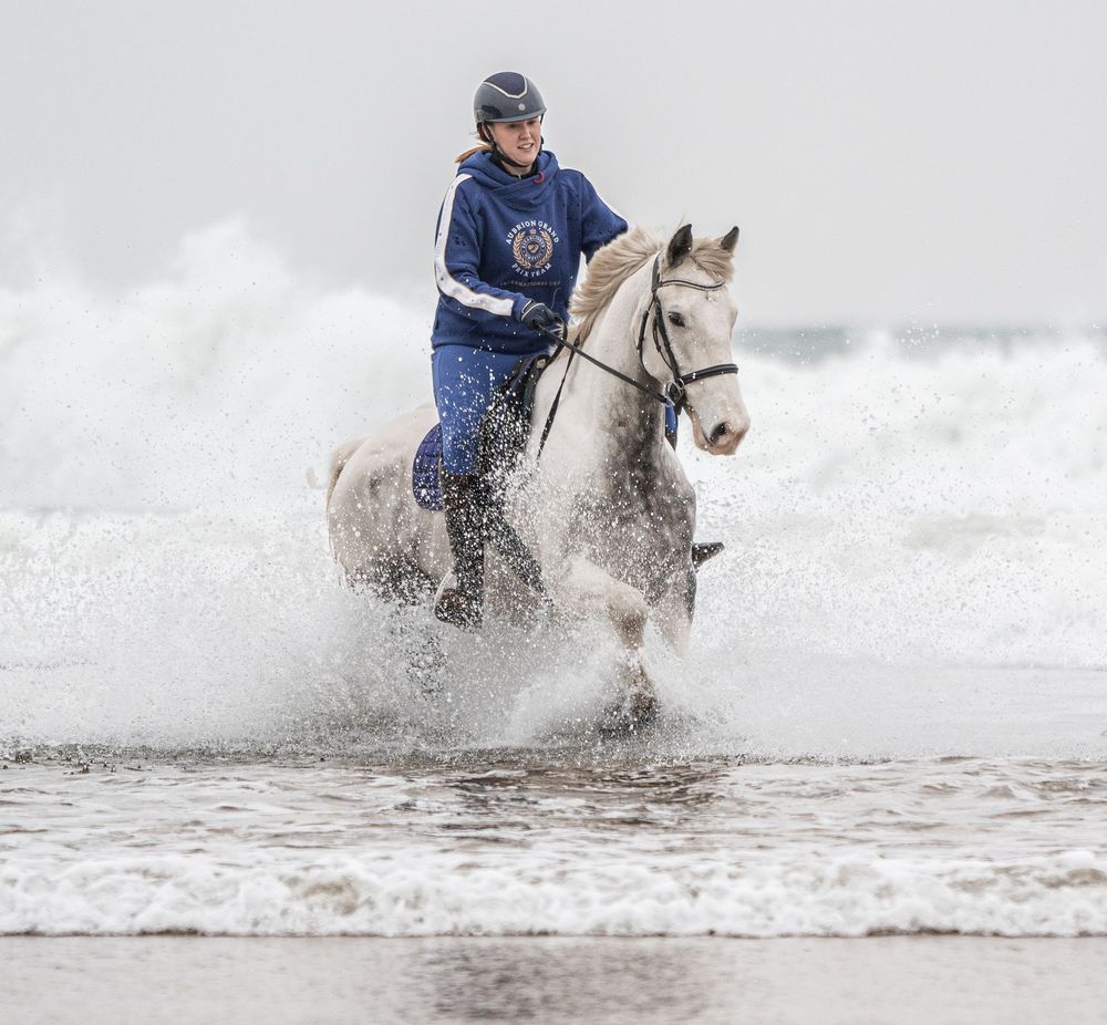 Rider on a white horse galloping through ocean waves on a misty beach creating dramatic splashes of water at Three Cliffs Bay.