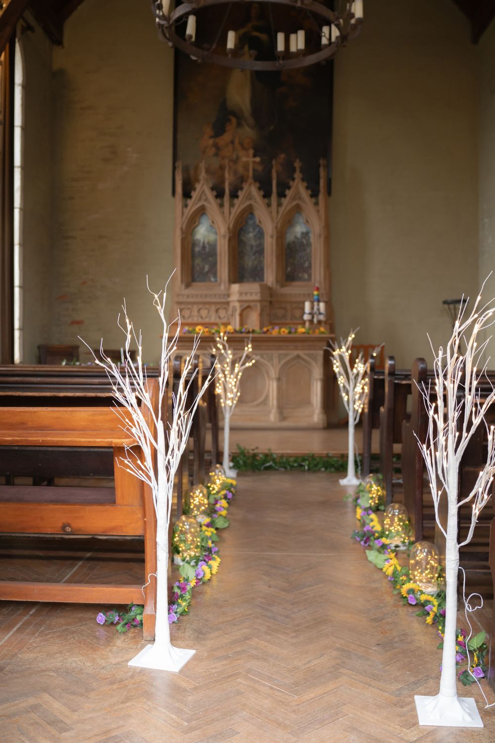 Illuminated white branch decorations line the aisle of a church with floral arrangements and wooden pews.