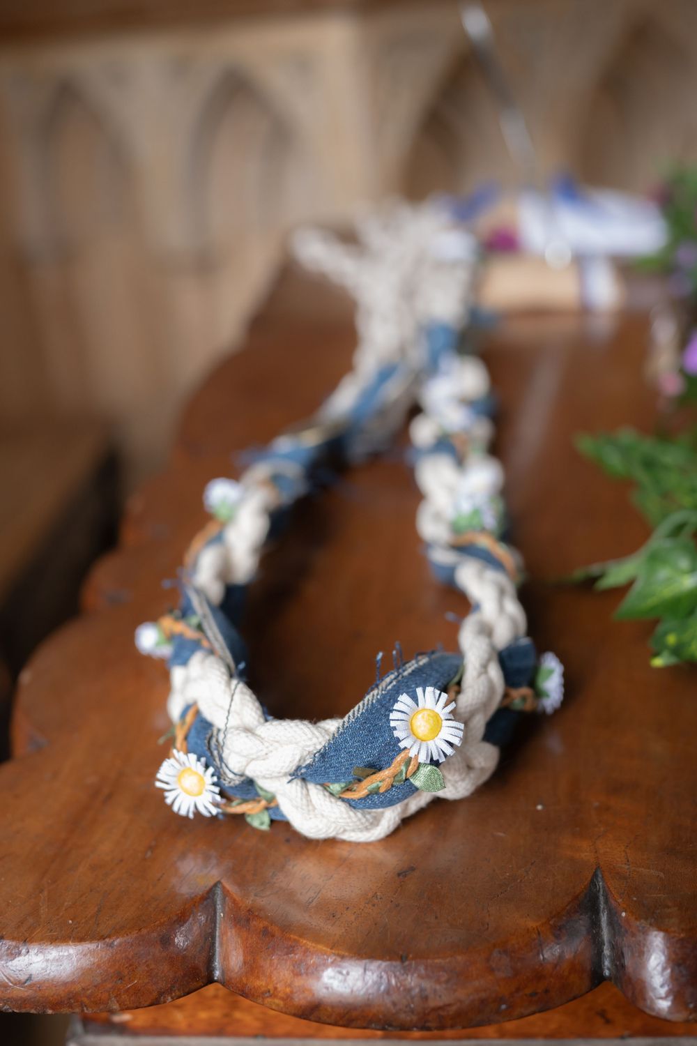 A rustic denim and lace braided bracelet with dainty white daisies lying on a wooden surface.