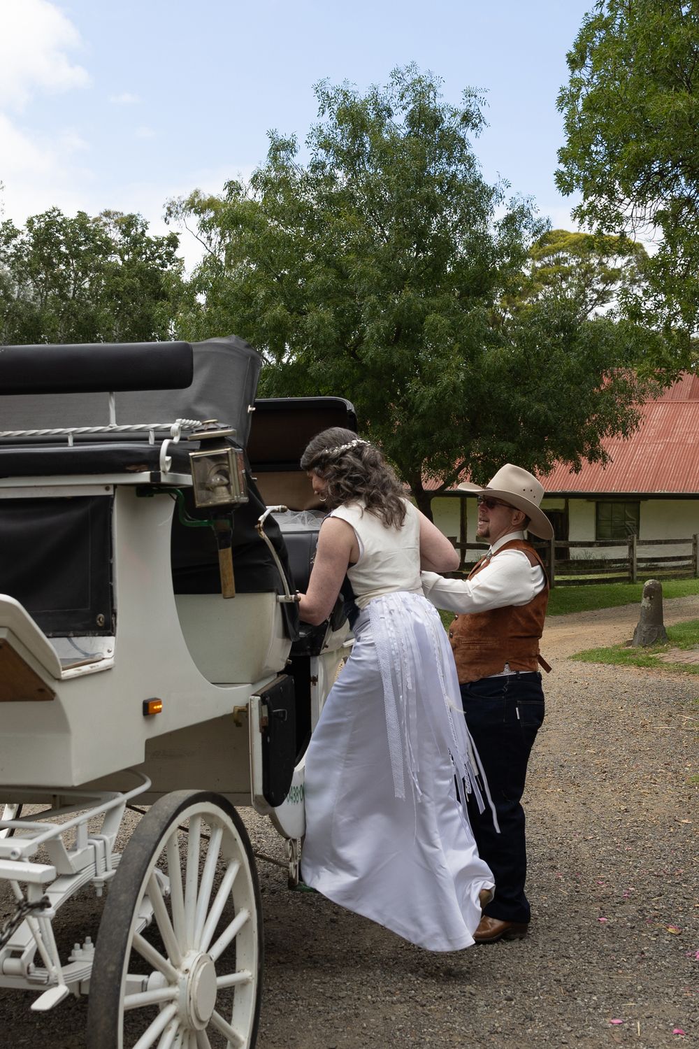 A white horse-drawn carriage is parked on gravel with people gathered around it on a sunny day.