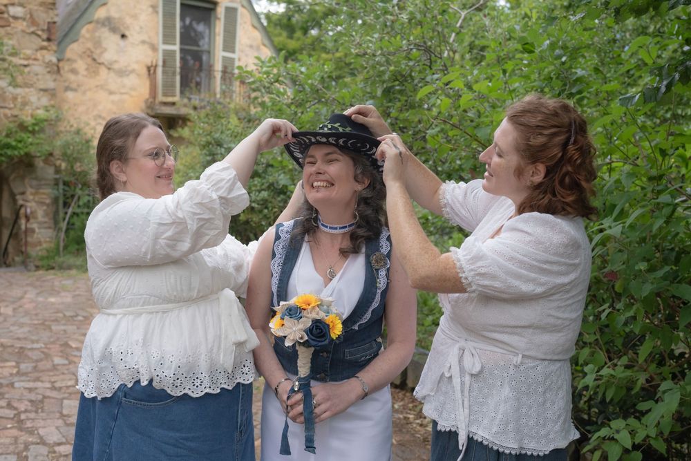 Three women in white lace and denim outfits share a joyful moment while placing a crown during an outdoor celebration.
