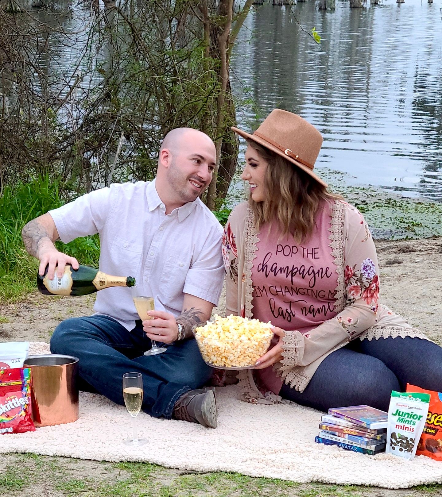 Engaged Couple enjoying a picnic by the water with snacks and blanket laid out.