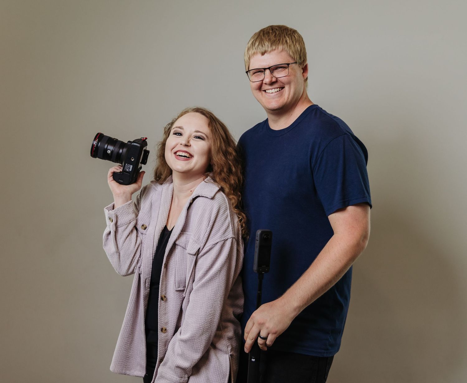 Two young people pose together against a neutral background, one wearing a red t-shirt and one in denim overalls.