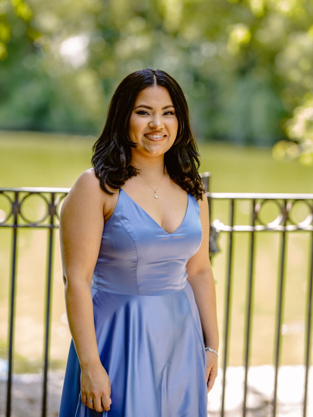 A woman in a periwinkle blue dress poses outdoors by a metal railing on a sunny day beautiful engagement shoot