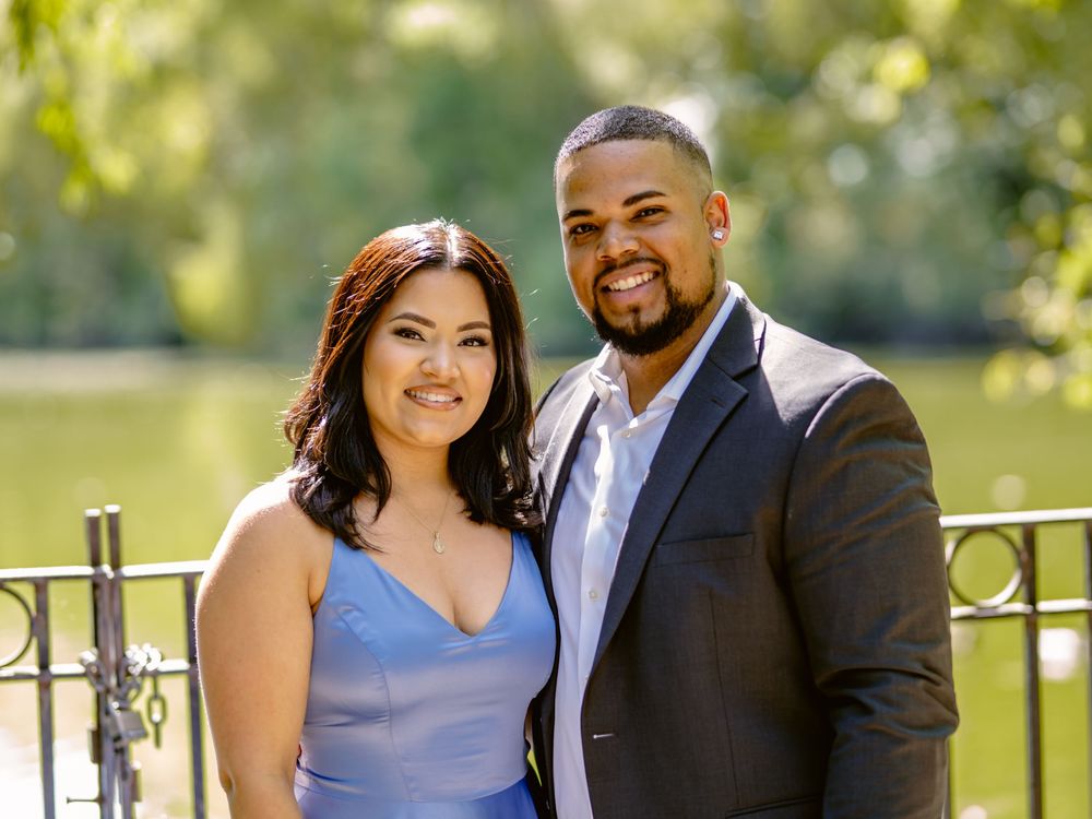 A couple in formal attire stands together by a railing in a garden setting on a sunny day beautiful engagement shoot