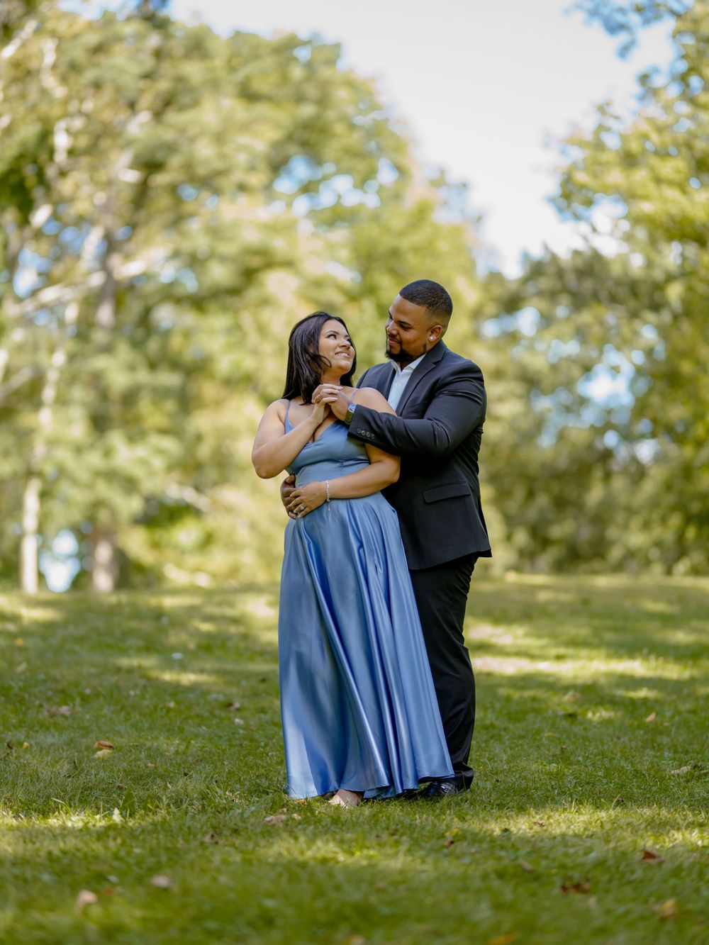 A couple shares an intimate embrace while standing in sunlit grass on a gentle hillside.
