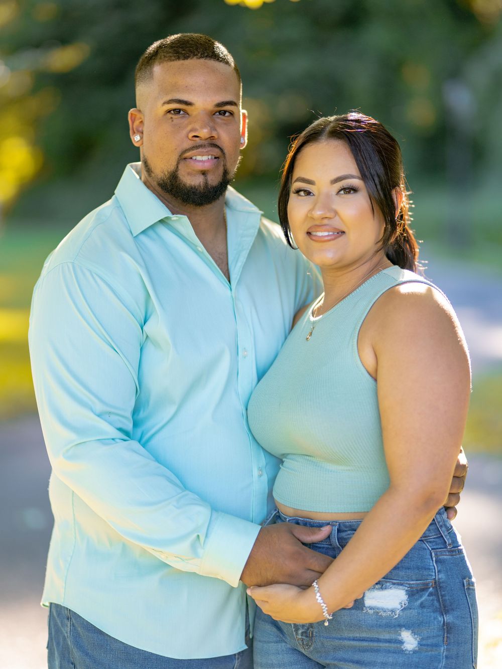 A couple poses outdoors in coordinating light blue outfits against a blurred nature background beautiful engagement shoot