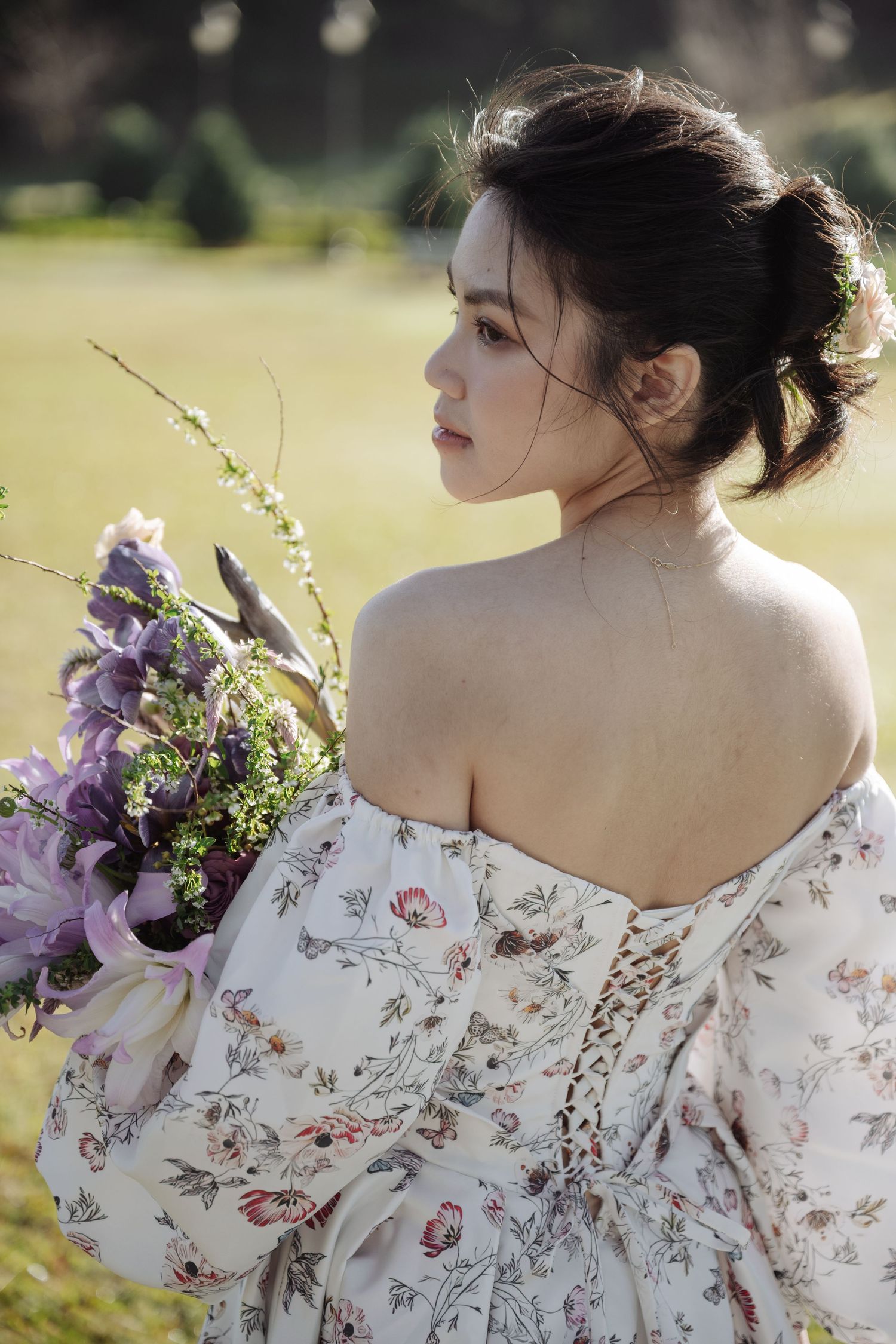 Young Asian woman in floral off-shoulder dress holding purple bouquet, viewed from behind in sunlit garden.