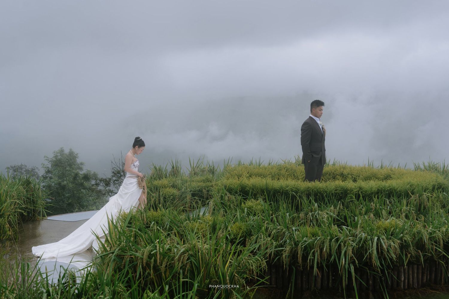 Dramatic misty landscape wedding photo of bride and groom standing apart on lush green rice terrace pathway.