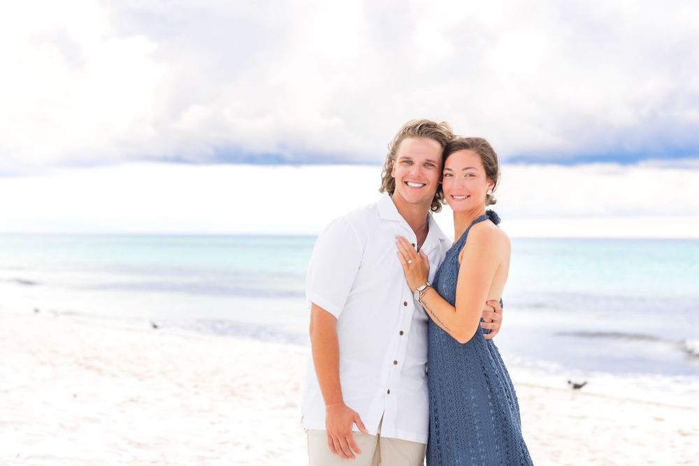 Two people embrace and laugh together by the ocean on a sunny beach wearing blue and white casual clothing.