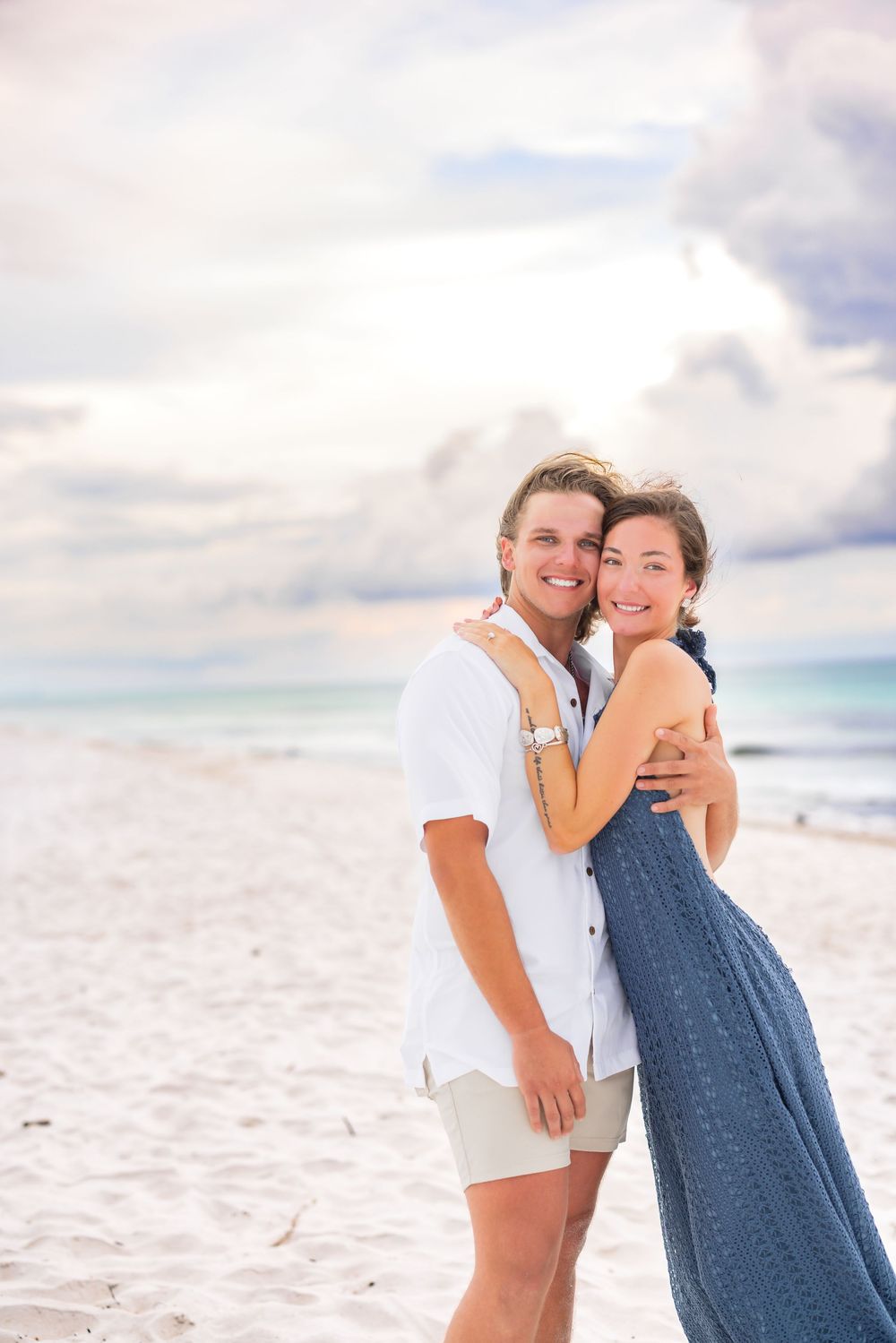 A couple embraces on a white sandy beach with cloudy skies as they pose for romantic sunset photos.