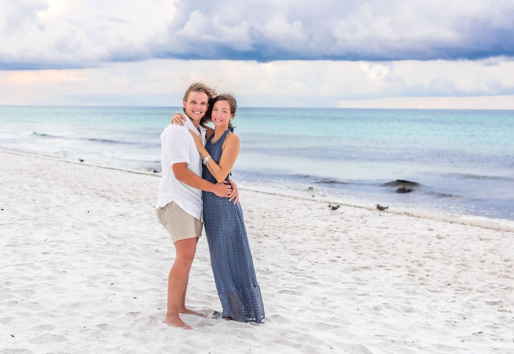 A couple poses romantically together on a white sandy beach with turquoise ocean waters and cloudy skies in view.