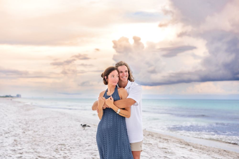 A couple embraces on a sandy beach at sunset with cloudy skies and ocean waves in the background.