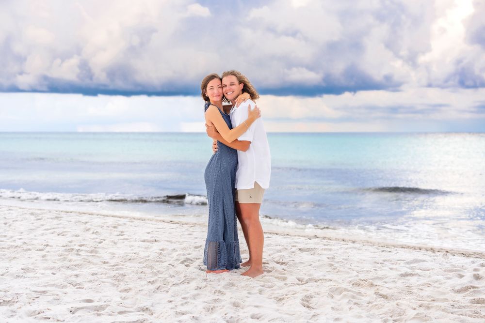 Two people embrace on a stunning white sandy beach with turquoise water and cloudy sky in the background.