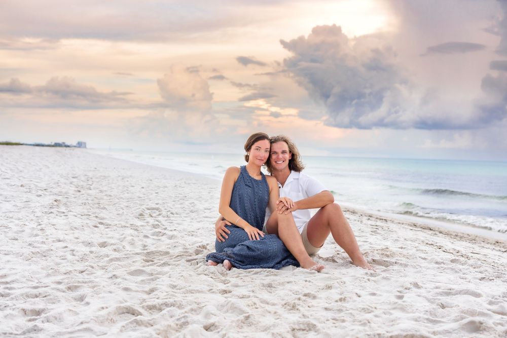 A romantic couple sitting on a white sandy beach at sunset enjoying each other's company.